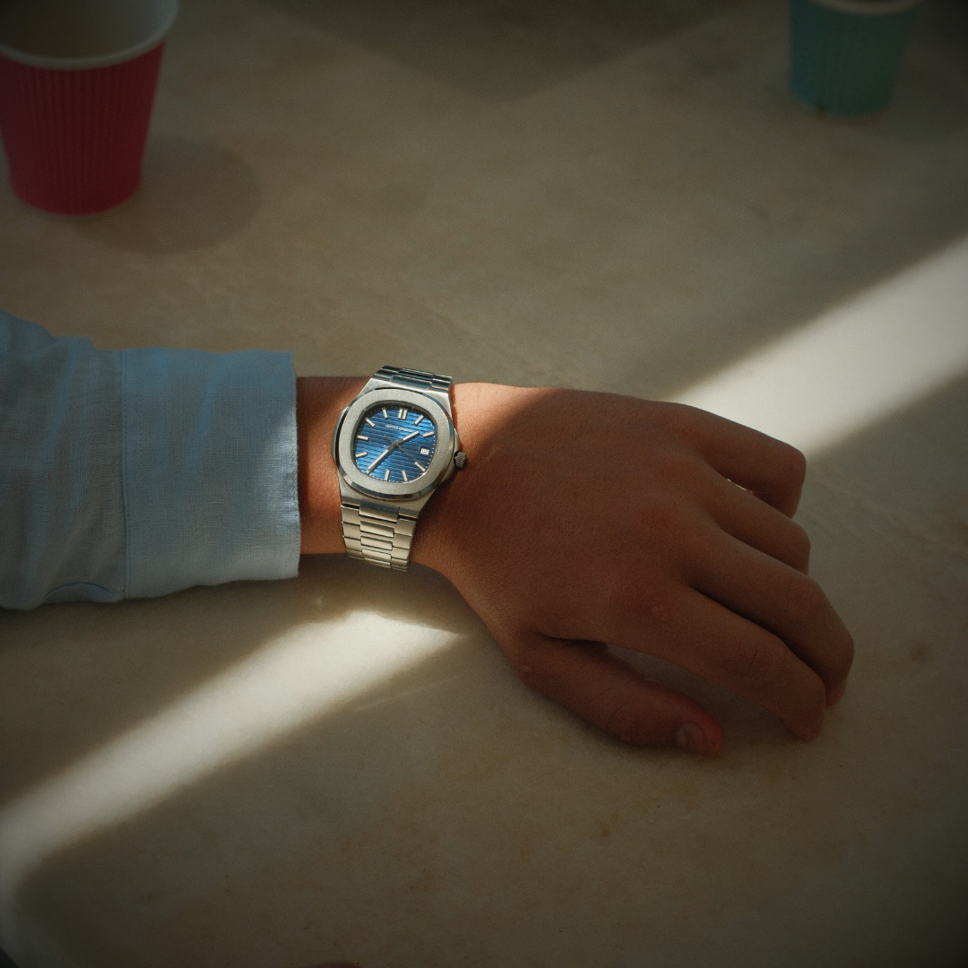 Hand wearing a silver watch with a blue face on a light surface.